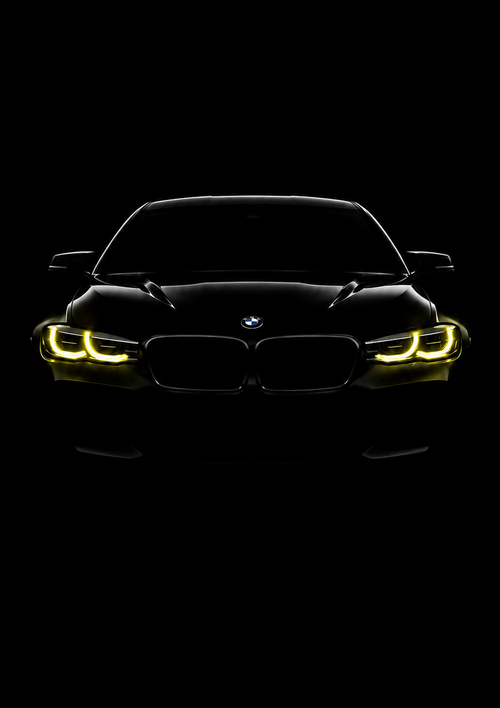 A framed black car poster featuring a front-facing BMW with glowing yellow headlights on a solid black background, presented in a sleek black frame.