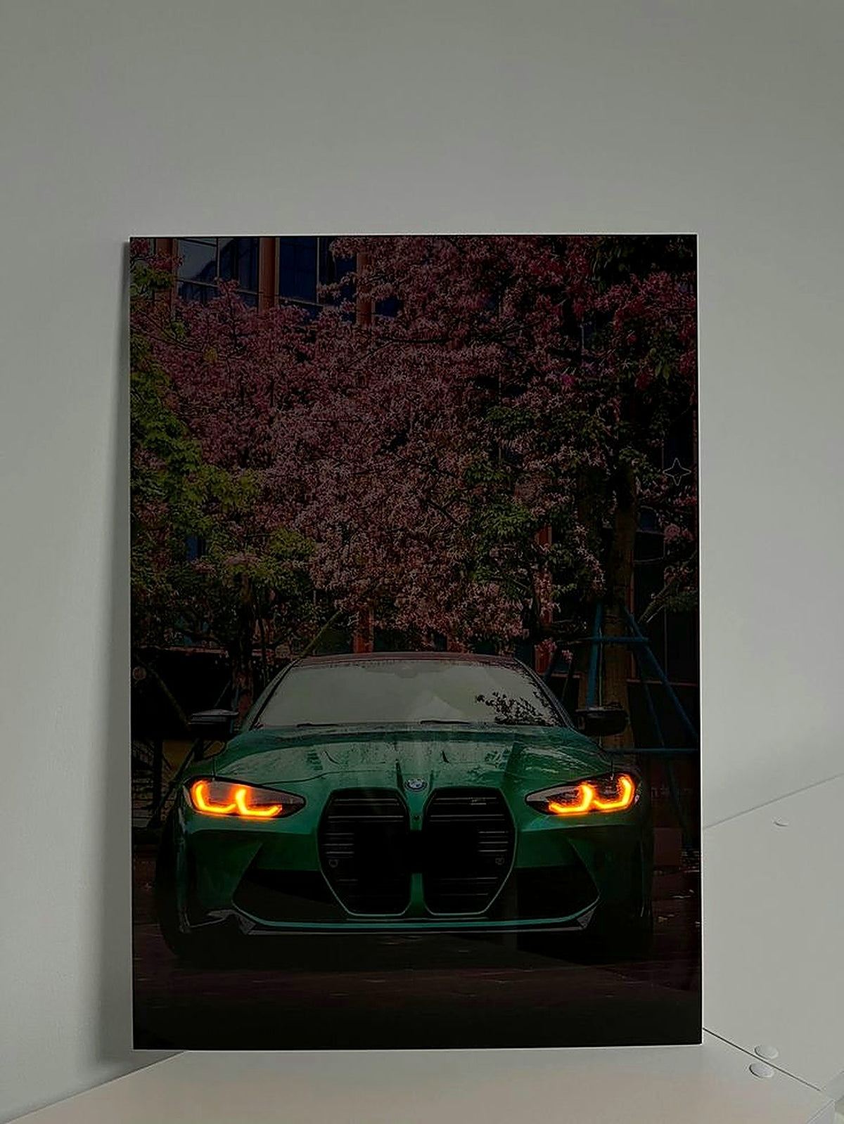 A vertical photographic print of a green BMW M2 with glowing amber headlights, set against a background of blooming pink cherry blossoms, displayed on a light gray wall.