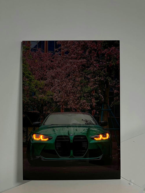 A vertical photographic print of a green BMW M2 with glowing amber headlights, set against a background of blooming pink cherry blossoms, displayed on a light gray wall.