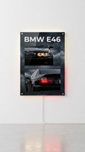 A vertical acrylic wall art print featuring the BMW E46, mounted on a white wall with a light strip underneath casting a red glow. The print shows two images of a black BMW E46 car against a dark cloudy sky, with 'BMW E46' written in white at the top.