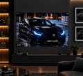 A large wall-mounted projection screen displays a black sports car on a wet city street at night. The room setting includes a dark wood console, framed wall art, a plant, and a sofa in a modern living room.