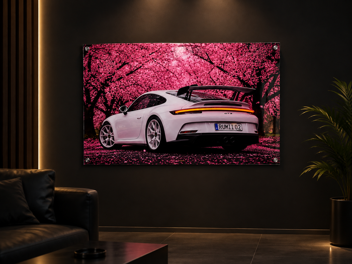 A large framed wall art print hanging on a dark wall in a modern living room. The artwork shows a white sports car from the rear, set against a vivid pink cherry blossom tree canopy with fallen petals on the ground.